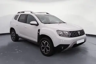 Dacia Duster blanc en 3/4 avant droit, avec calandre noire et jantes alliage, stationné sur un sol gravillonné.